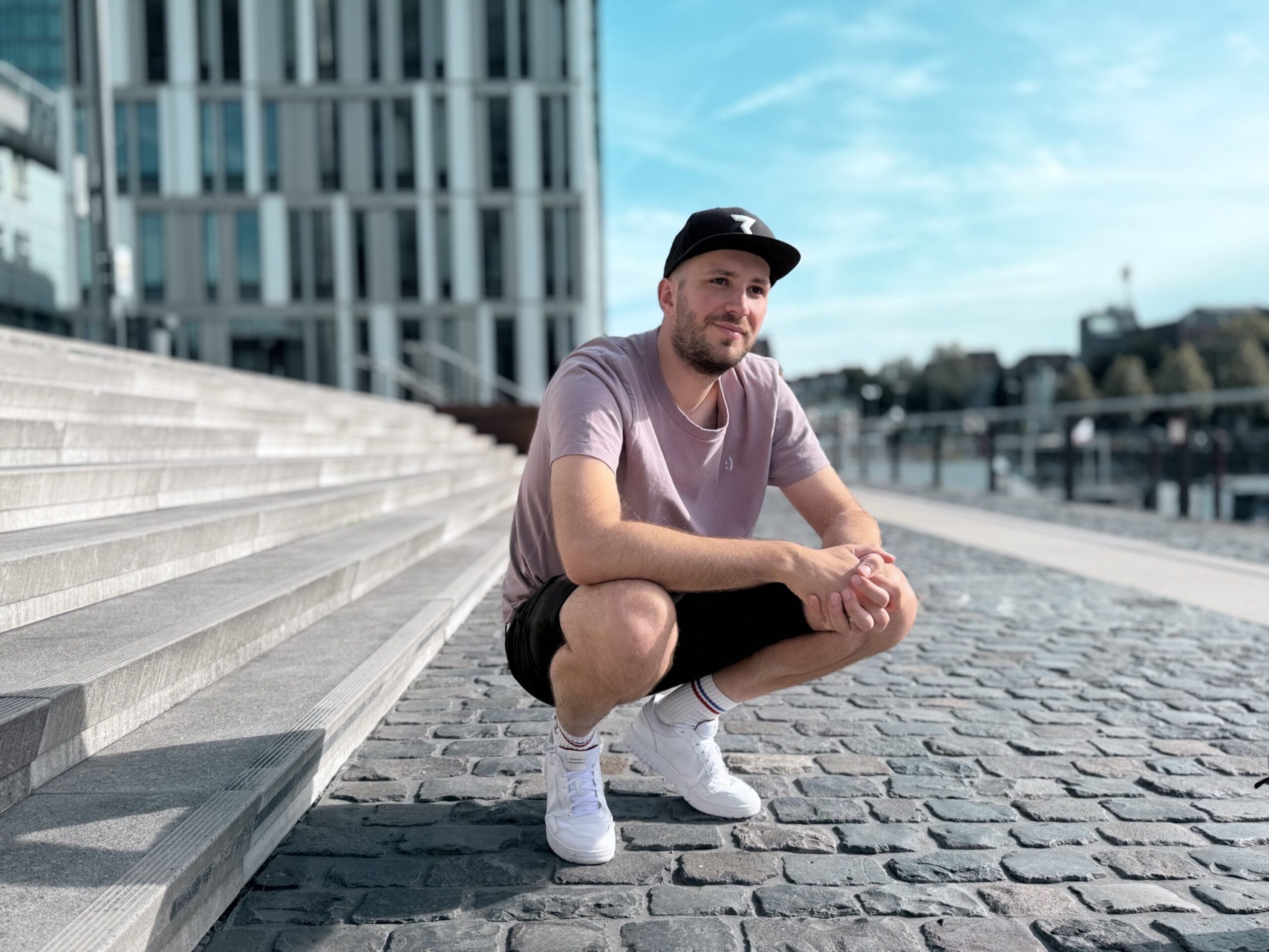 Junger Mann mit Vollbart und schwarzer Baseball-Cap, der lässig kauernd neben breiten, grauen Beton-Treppenstufen sitzt. Hellrosa T-Shirt, schwarze Shorts, helle Socken und weiße Sneakers; die Pose wirkt entspannt und selbstbewusst, mit Händen auf den Knien und leichtem Lächeln. Im Hintergrund dominiert eine moderne Fassade eines Hochhauses, flankiert von Kopfsteinpflaster, einer Hafenpromenade mit Wasser und klarem blauen Himmel.